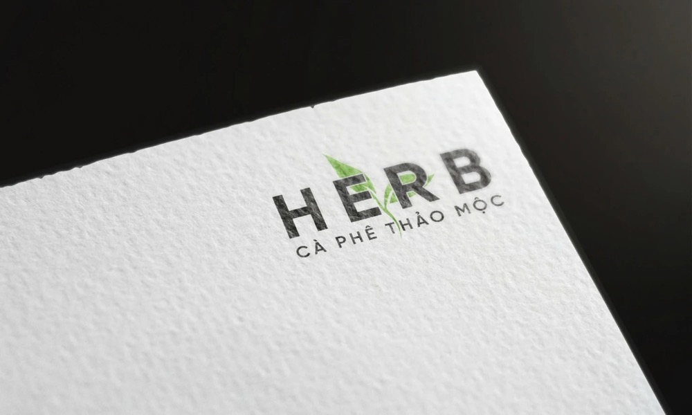Logo HERB - CÀ PHÊ THẢO MỘC chữ xám đen và chiếc lá xanh in trên giấy trắng có vân