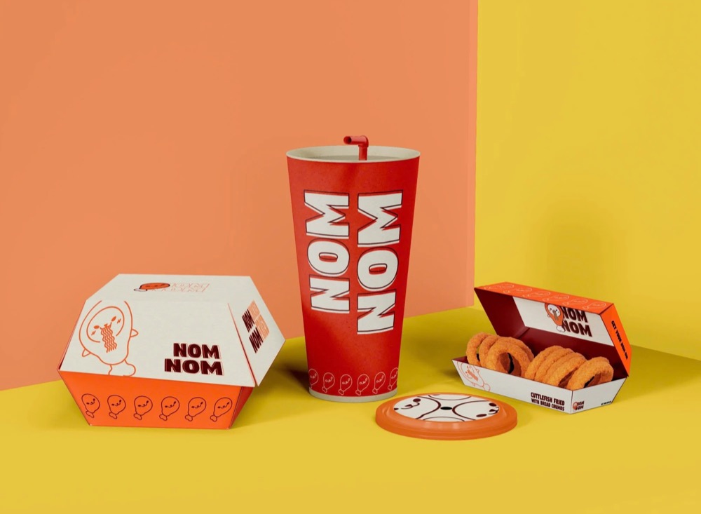 Bộ bao bì NOM NOM gồm hộp burger màu kem và cam, ly giấy đỏ in chữ NOM NOM lớn, nắp ly và hộp đồ chiên mở nắp trên nền cam vàng
