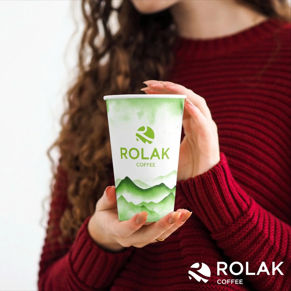 Người cầm ly giấy ROLAK COFFEE màu trắng xanh lá với logo và họa tiết núi màu nước