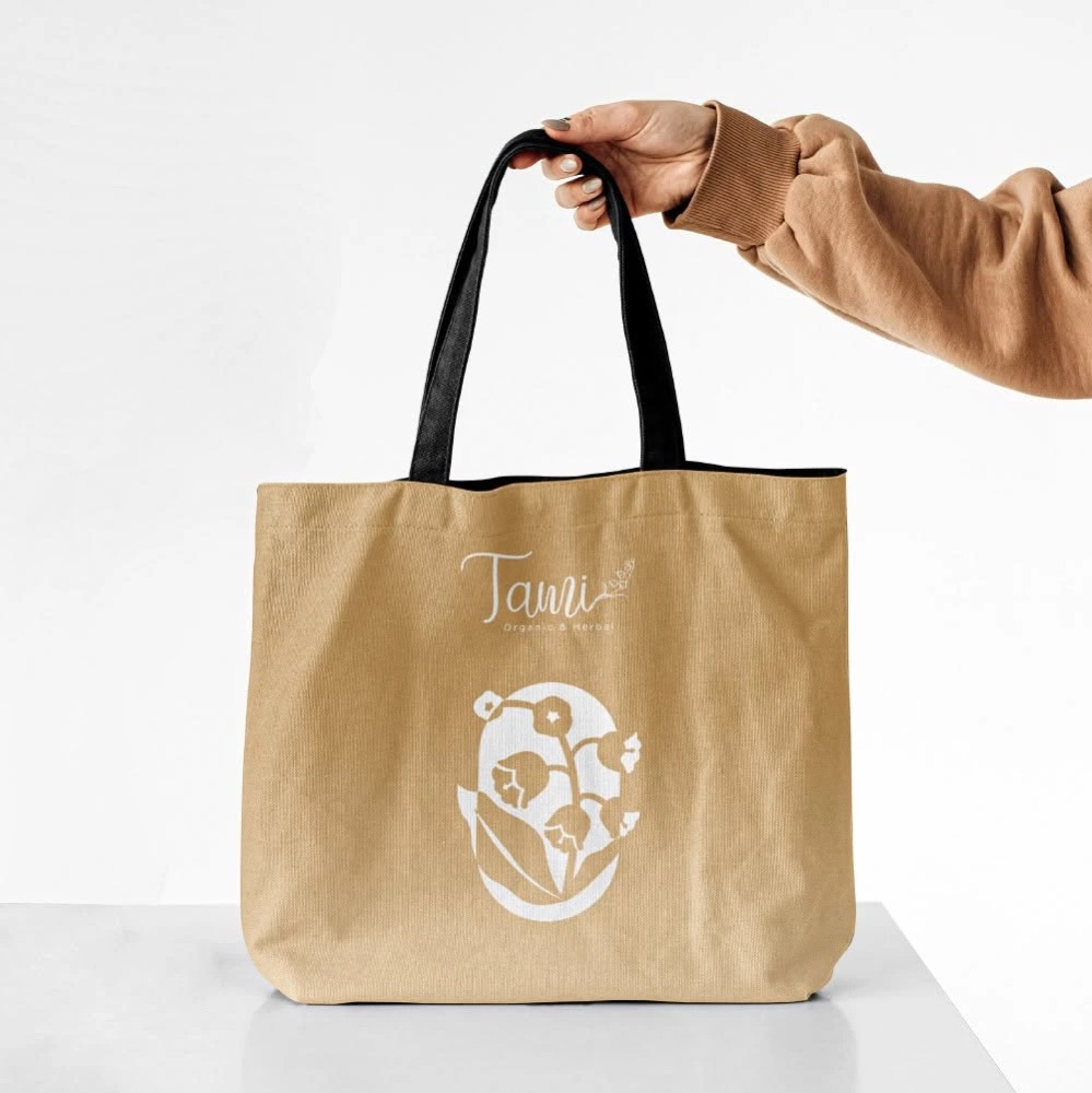 Túi tote vải màu nâu be quai đen in logo TAMI Organic & Herbal và biểu tượng hoa linh lan màu trắng