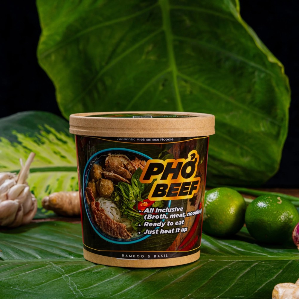 Một ly Phở Beef BAMBOO & BASIL đặt trên lá xanh cùng chanh, gừng và tỏi, tem thân ghi All inclusive, Ready to eat, Just heat it up